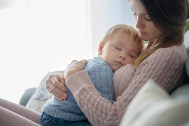 Onu evde, sarılma onun hasta yürümeye başlayan çocuk tutan genç anne, güneşli oturma odası