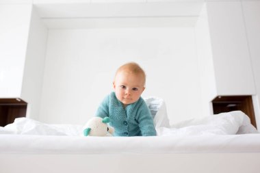 Mutlu bir şekilde beyaz güneşli, parlak yatak odasında kamera gülümseyen tutmak örme oyuncak, küçük şirin bebek çocuk, çocuk örme kazaklı,