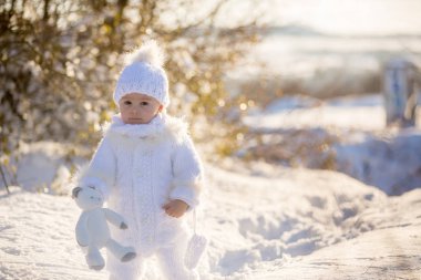 Bebek kışın karda Teddy ile oynuyor. El yapımı beyaz kar elbisesi içinde küçük bir çocuk, gün batımında oyuncak ayıyı tutuyor, kış parkında açık havada oynuyor. Çocuklar karlı parkta oynuyor.
