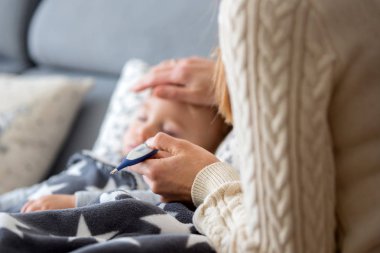 Hasta çocuk, çocuk oturma odasında kanepede yatıyor, ateşi var, annesi ateşini ölçüyor, evde dinleniyor.