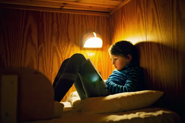 バック グラウンドでライトとベッドに横たわって本を読んで楽しんで面白い表情のかわいい小さな幼児男の子 夕方ホーム余暇 本の魔法の世界 — ストック写真