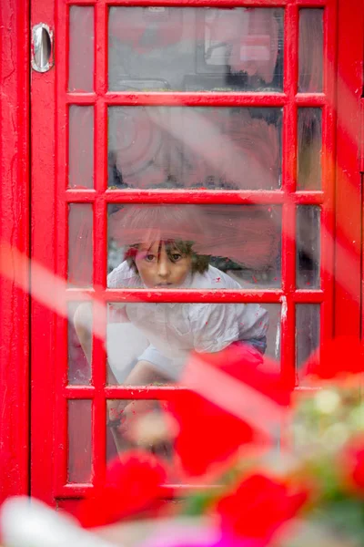 Χαριτωμένο αγόρι, παιδί, καλώντας από ένα κόκκινο τηλεφωνικό θάλαμο στην πόλη — Φωτογραφία Αρχείου
