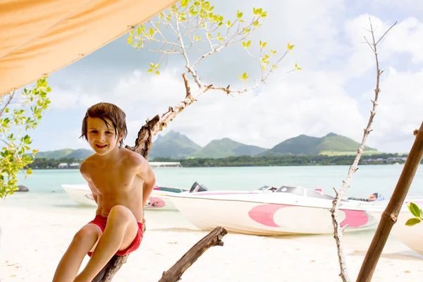Słodki chłopiec w wieku przedszkolnym, siedzący na gałęzi drzewa na plaży — Zdjęcie stockowe