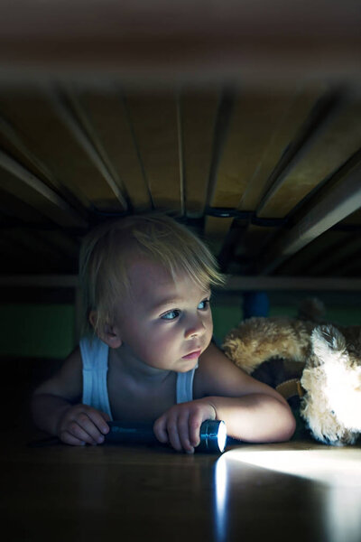 Little child, hiding under the bed, hugging teddy bear and holdi