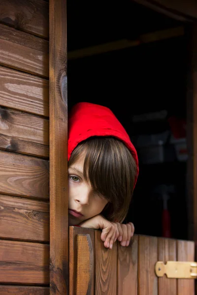 Niño preadolescente en sudadera roja, escondido detrás de una puerta de madera, mira — Foto de Stock