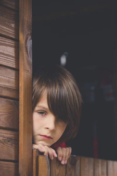 Niño preadolescente en sudadera roja, escondido detrás de una puerta de madera, mira —  Fotos de Stock