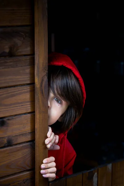Niño preadolescente en sudadera roja, escondido detrás de una puerta de madera, mira — Foto de Stock