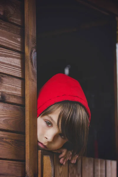 Niño preadolescente en sudadera roja, escondido detrás de una puerta de madera, mira —  Fotos de Stock