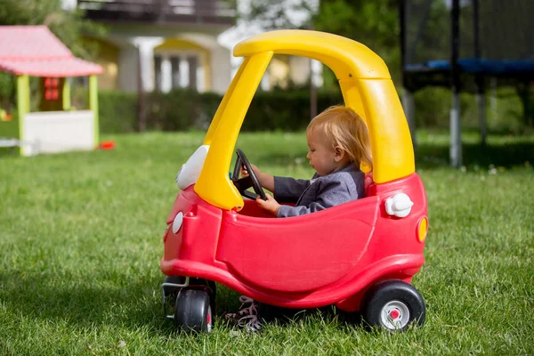 Lindo niño pequeño, montar gran juguete de plástico coche rojo en el parque —  Fotos de Stock