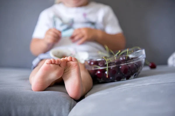 Sweet toddler boy, sitting on a couch, eating cherries — Stock Photo, Image