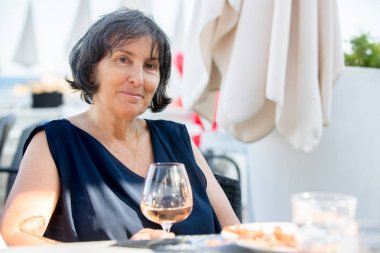 Yaşlı kadın, sahilde bir bardak gül şarabı içiyor 