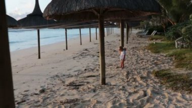 Küçük yürümeye başlayan çocuk, gün doğumunda sahilde oyuncak ayı ile oynarken, plajda Şemsiyeler