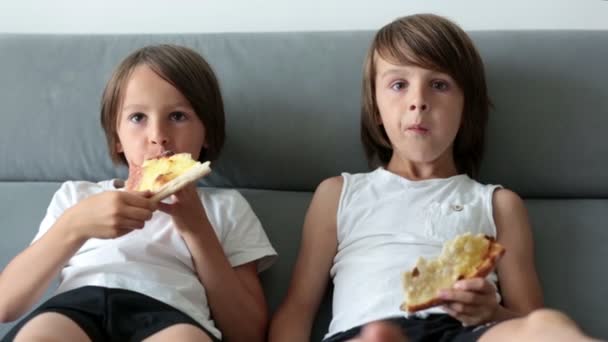 かわいい子供たちは ソファに座って ピザを食べてテレビを見ています 家でピザパーティーの日にピザから一口を取って空腹の子供 — ストック動画