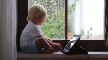 Sevimli küçük çocuk, pencere kalkanı üzerinde oturan, yağmurlu bir gün yaz tablet üzerinde oynuyor
