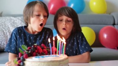 Beautiful  boy in blue shirt, celebrating his birthday, blowing candles on homemade baked cake, indoors. Birthday party for kids