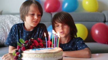 Beautiful  boy in blue shirt, celebrating his birthday, blowing candles on homemade baked cake, indoors. Birthday party for kids