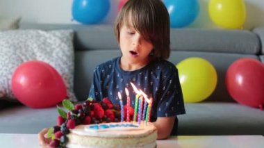 Beautiful  boy in blue shirt, celebrating his birthday, blowing candles on homemade baked cake, indoors. Birthday party for kids