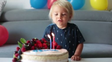 Üç yaşında mavi gömlekli güzel bir çocuk. Doğum gününü kutluyor. Ev yapımı pastaya mum üflüyor. Çocuklar için doğum günü partisi
