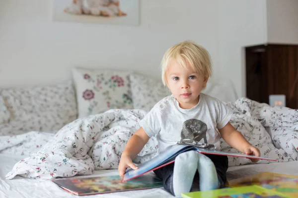 可爱的蹒跚学步的男孩，在家里看书，坐在床上，很多很多 — 图库照片