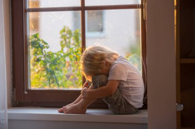 Terk edilmiş küçük çocuk, pencerenin camına oturmuş, gün batımını seyrediyor.