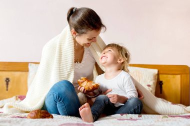 Anne ve çocuk, birlikte eğleniyor, kitap okuyor ve yatakta kruvasan yiyorlar.