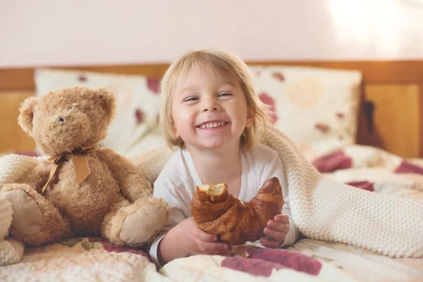 かわいいブロンドの幼児の子供 本を読んで ベッドでクロワッサンを食べます 喜んで笑って — ストック写真