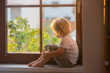 Terk edilmiş küçük çocuk, pencerenin camına oturmuş, gün batımını seyrediyor.