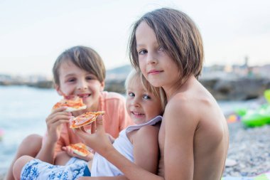 Tatlı çocuk, kumsalda pizza yiyor, eğleniyor, mutlu bir şekilde gülümsüyor, çocuk sahilde yemeğin tadını çıkarıyor.