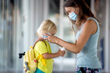 Anne ve çocuk, küçük çocuk, yüz maskesi takıyor ve havaalanında dezenfeksiyon kullanıyor, bir uçuşa hazırlanıyor, tüm önlemlerle aile gezisi.