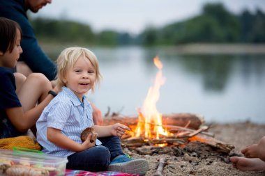 Küçük çocuk, çocuk ailesiyle piknik yapıyor ve yaz akşamları nehir kenarında kamp ateşi yakıyor.