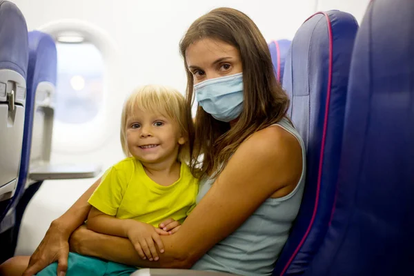 Mother and child, boy and mom, wearing masks and using disinfection, sitting in airplane at the airport, preparing for a flight, family trip with all precautions