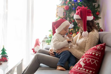 Güzel genç anne ve sarışın çocuk, rahat bir evde oturuyor, Noel için süslenmiş, kitap okuyor, sarılıyor ve öpüşüyor.