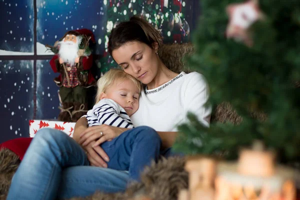 Güzel genç anne ve sarışın çocuk, rahat bir oturma odasında oturuyor, Noel için süslenmiş, sarılıyor ve öpüşüyor.