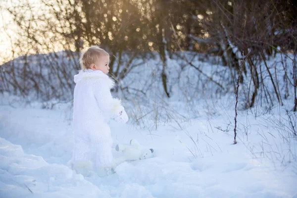 雪の中でテディと遊んで赤ちゃん 冬時間 手作りの白いスノースーツの小さな幼児の少年は 日没にテディベアを持ち 冬の公園で屋外で遊んでいます 雪の公園で遊ぶ子供たち — ストック写真