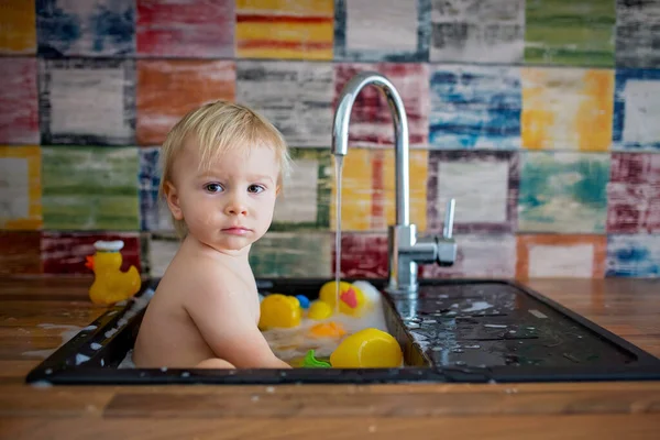 Cute smiling baby taking bath in kitchen sink. Child playing with foam and soap bubbles in sunny kitchen with rubber ducks and toys. Little boy bathing, fun with water
