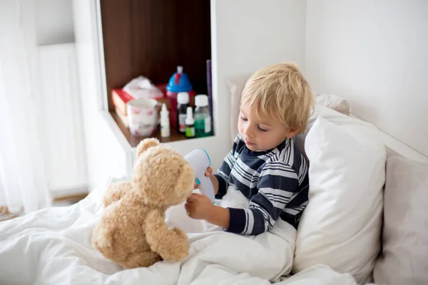 小孩子金发碧眼 躺在床上和泰迪熊玩耍 生病时 用数字温度计检查体温 — 图库照片