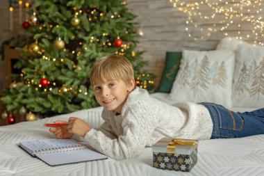 Sevimli anaokulu çocuğu, şöminenin önünde oturuyor, Noel hediyesini tutuyor ve evde kitap, eğlence ve mutluluk okuyor.