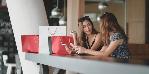 Deux belles femmes asiatiques vérifiant leur smartphone et bavardant ensemble après avoir fait du shopping dans le centre commercial — Photo