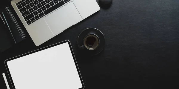 Top view of dark workplace with blank screen tablet and laptop computer on black leather background