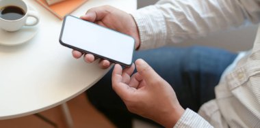 Boş ekranlı akıllı telefonu kahve fincanıyla rahat çalışma alanında tutan işadamının yakın görüntüsü 