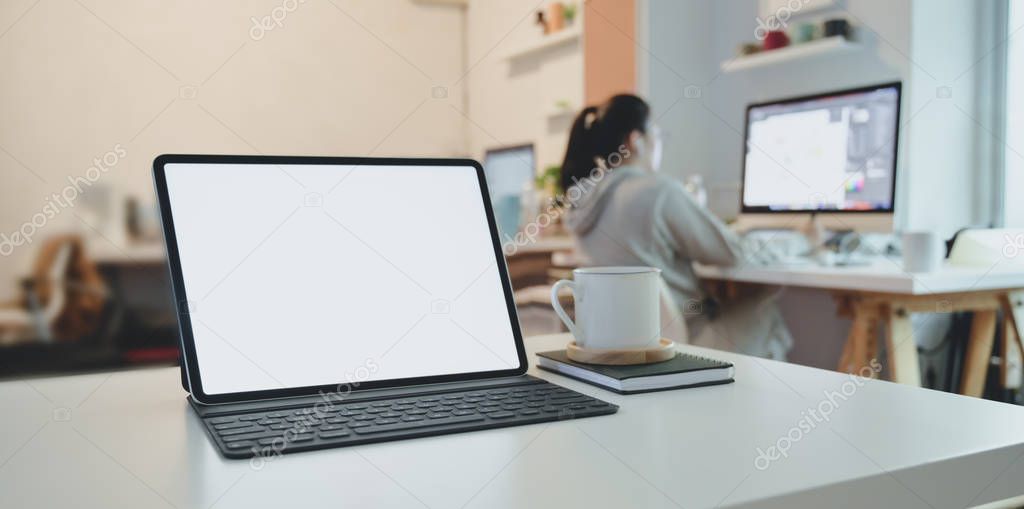 Blank screen tablet with keyboard on white wooden table with office supplies with businesswoman in the background 