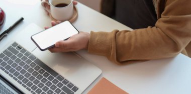 Genç adam elinde boş ekran akıllı telefonu tutarken dizüstü bilgisayarda çalışıyor ve kahve içiyor. 