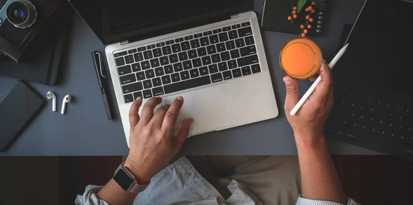 Visão superior do homem trabalhando em seu projeto com computador portátil enquanto bebe um copo de suco de laranja no espaço de trabalho moderno escuro — Fotografia de Stock