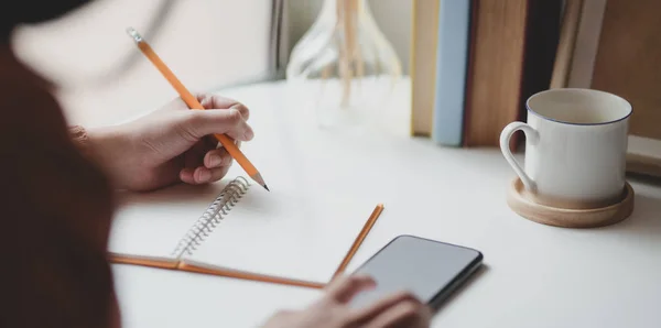 Vista de cerca de la joven mujer escribiendo su idea en el cuaderno mientras toca su teléfono inteligente en la cómoda habitación de la oficina — Foto de Stock