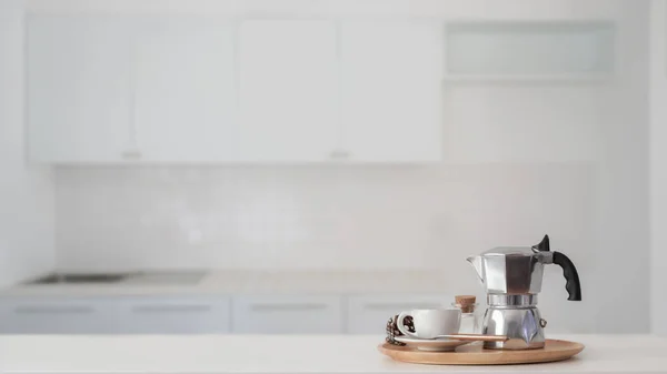 Coffee cup and Moka pot in wooden tray on white counter with blurred kitchen in the background — Stock Photo, Image