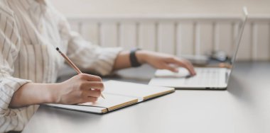 Laptop bilgisayarda yazı yazan ve ofis odasında deftere yazan tutkulu genç iş kadınının yakın plan görüntüsü. 