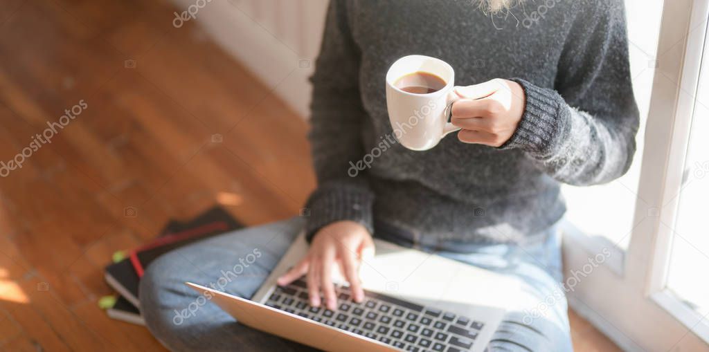 Close-up view of young freelancer working on her project and drinking hot cocoa while sitting near the windows at her workplace 