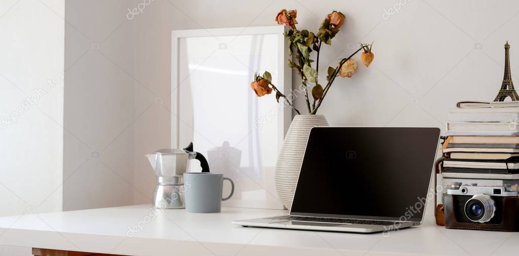 Minimal workspace with laptop computer, camera, office supplies and dry roses vase on white table 