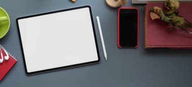 Boş ekran tableti, ofis gereçleri ve koyu gri masa arkaplanlı akıllı telefonu olan modern işyerinin üst görüntüsü 