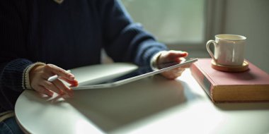 Beyaz sehpanın üzerinde dijital tablet kullanan bir kadının, oturma odasında kitap ve fincanla çekilmiş bir fotoğrafı. 